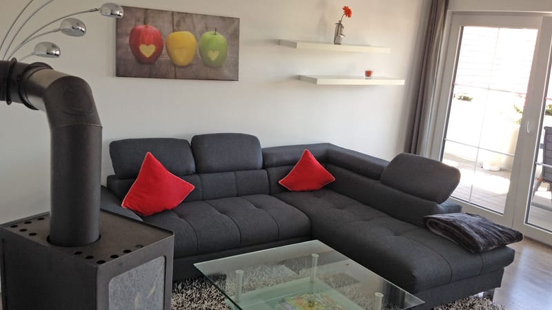 Wohnbereich mit dunkelgrauem Sofa, roten Kissen und schwarzem Ofen, rechts eine Glastür zum Balkon