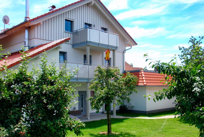 Das Haus von der Gartenseite mit Balkonen und rotem Ziegeldach, davor Rasen und Obstbäume